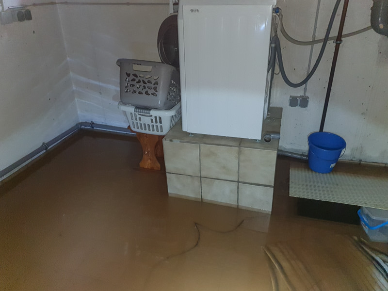 In einem Waschkeller steht eine Waschmaschine auf einem Podest. Rechts davon ist der Pumpensumpf. Der Boden ist bedeckt mit schlammigen Wasser der Flut.